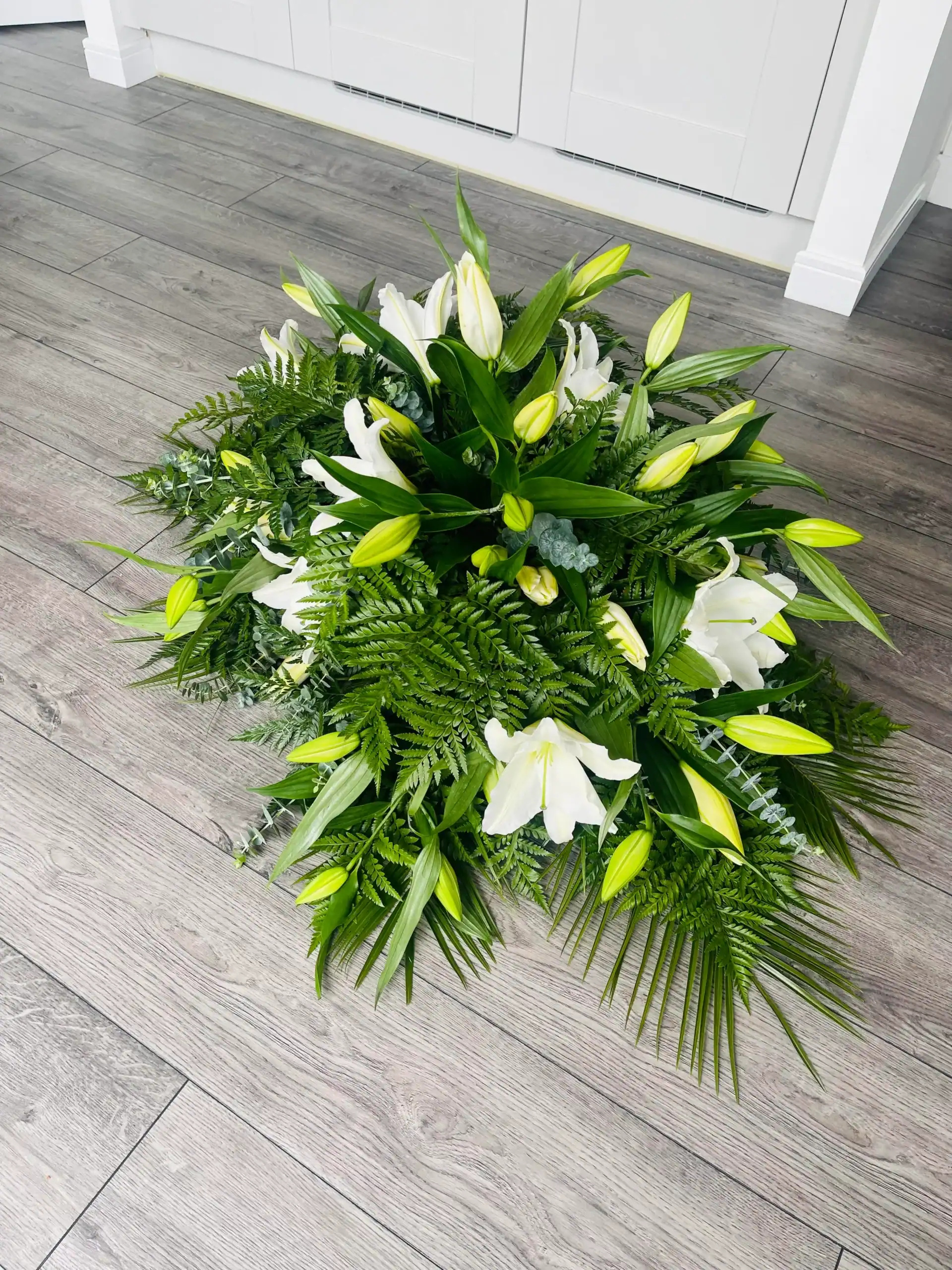 Elegant white lily floral arrangement with lush greenery on wooden floor.